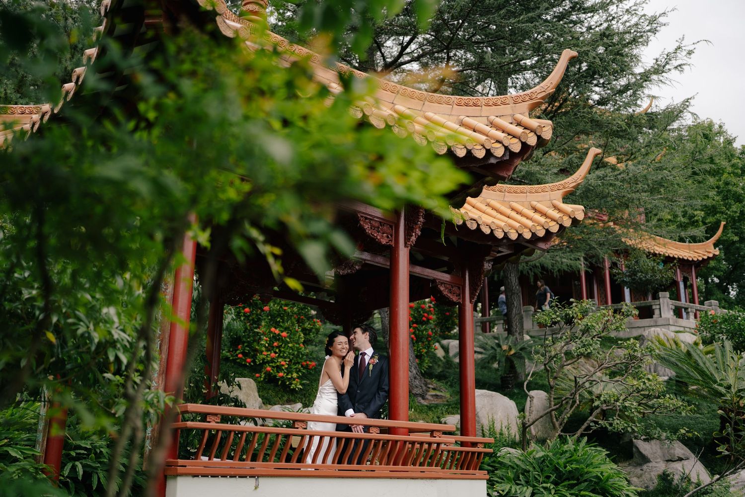 Couple in wedding attire embracing in a traditional Chinese red pavilion surrounded by lush greenery.