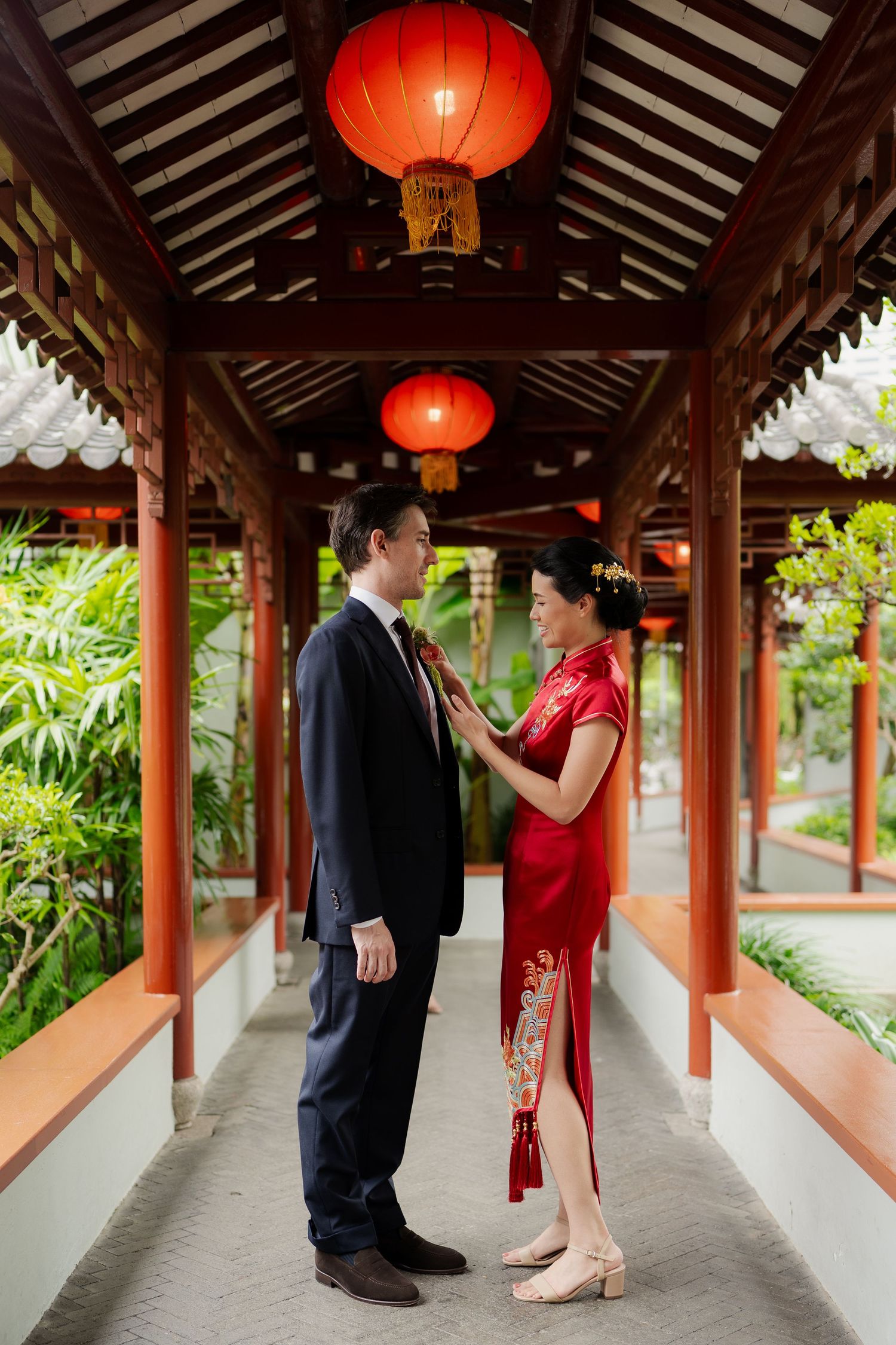 Couple in traditional Chinese wedding attire standing under red lanterns in an elegant wooden pavilion.