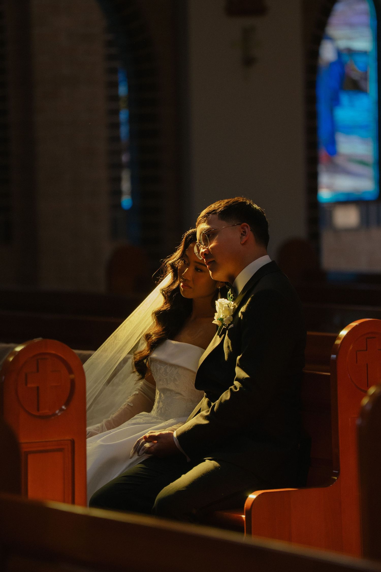 Bride and groom share an intimate moment seated in wooden church pews, with stained glass windows glowing softly behind them.
