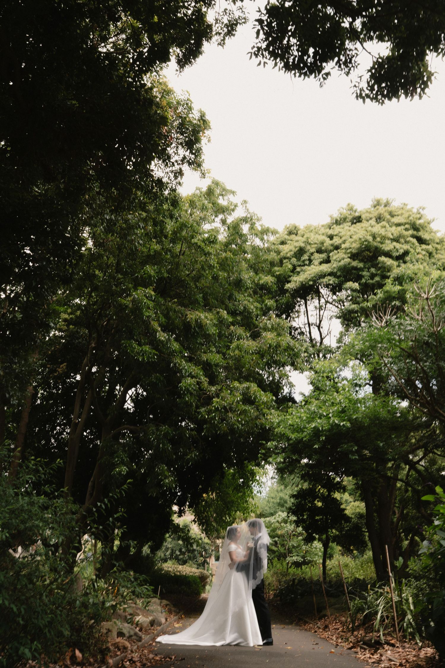 A bride and groom share a romantic moment on a garden path surrounded by lush green trees on their wedding day.