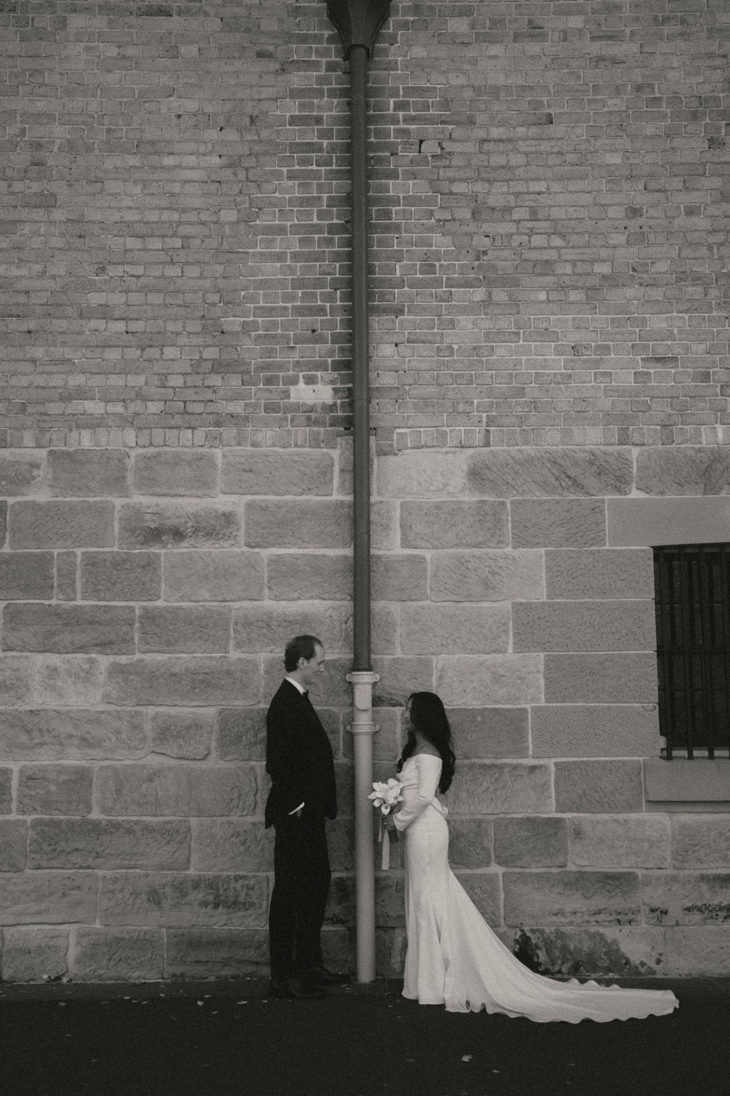 Black and white wedding photo of bride and groom standing on either side of a drainpipe against a stone brick wall.