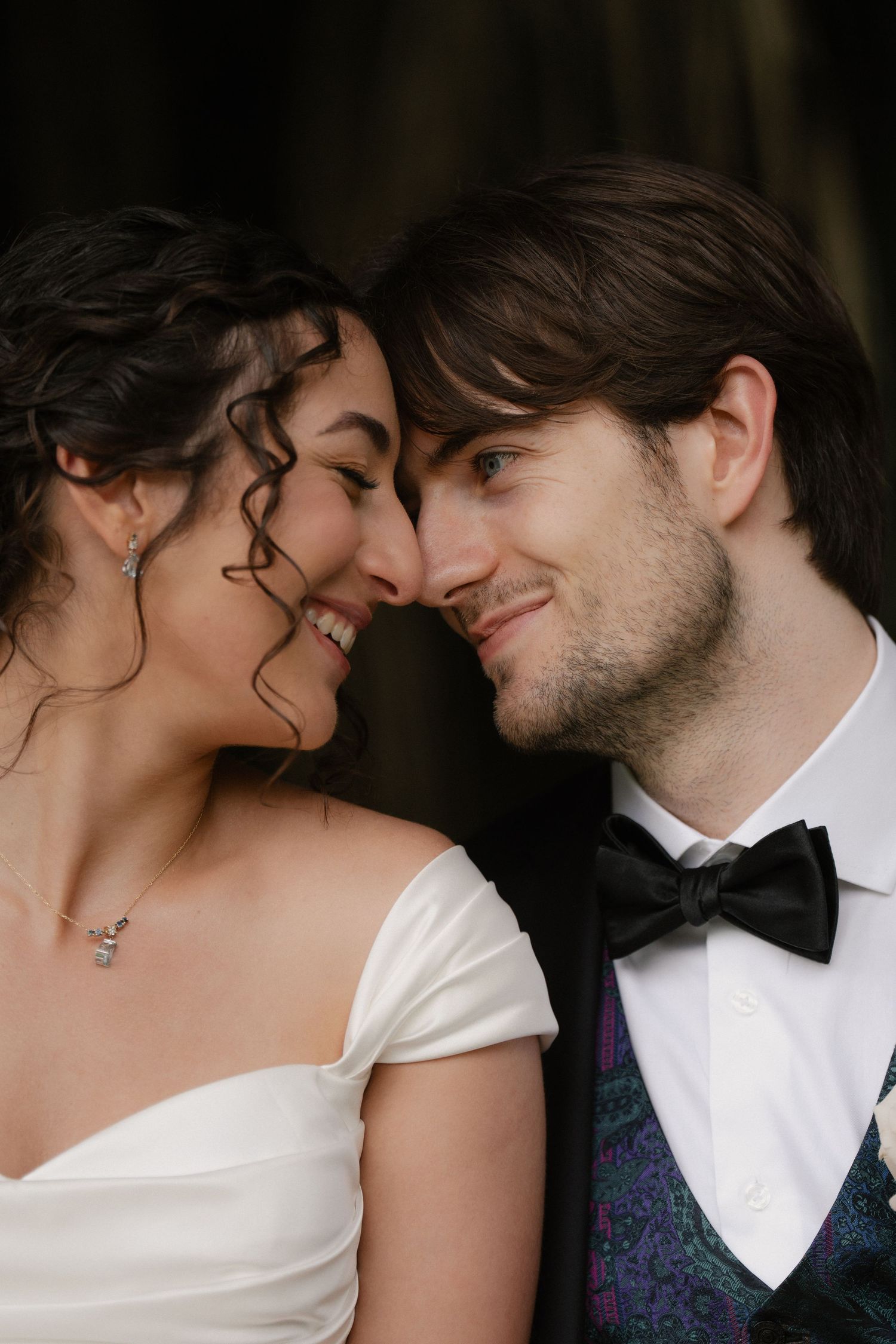 A smiling bride and groom in formal wedding attire share a romantic nose-to-nose moment, radiating joy and love.