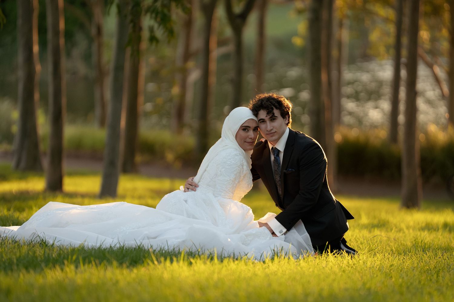 Muslim bride in white hijab and gown sits with groom in black suit on green grass in a sunlit park.