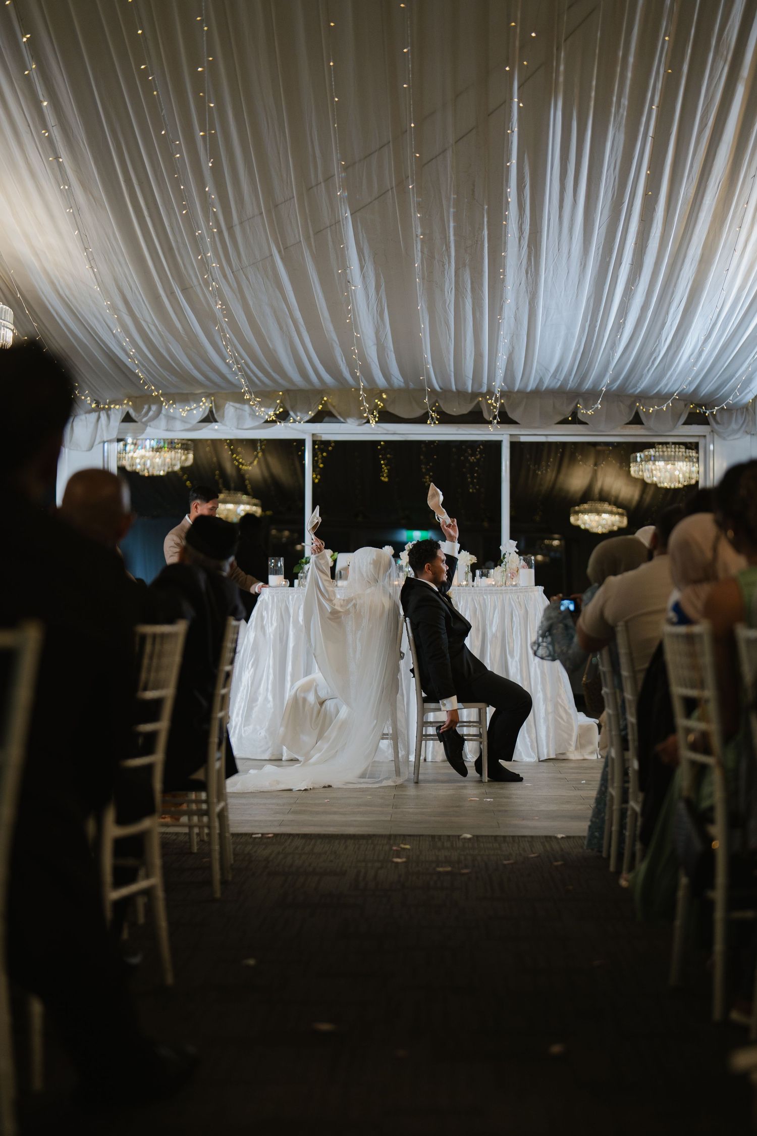 Elegant wedding reception in a white draped tent venue with fairy lights, guests seated in chiavari chairs as groom celebrates.