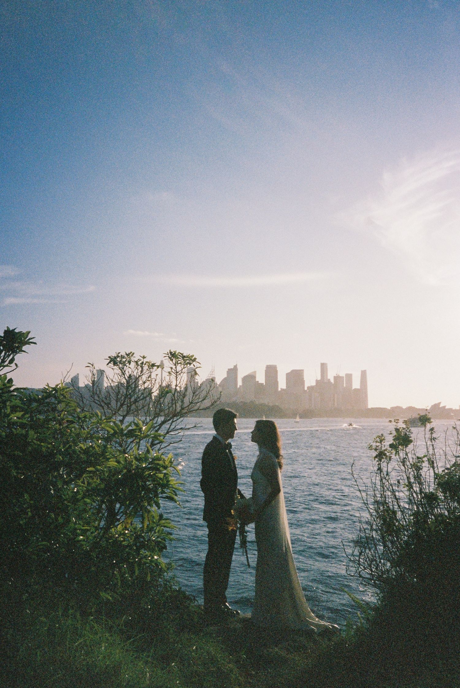 Couple in wedding attire holding hands by waterfront with city skyline silhouetted against golden sunset sky.