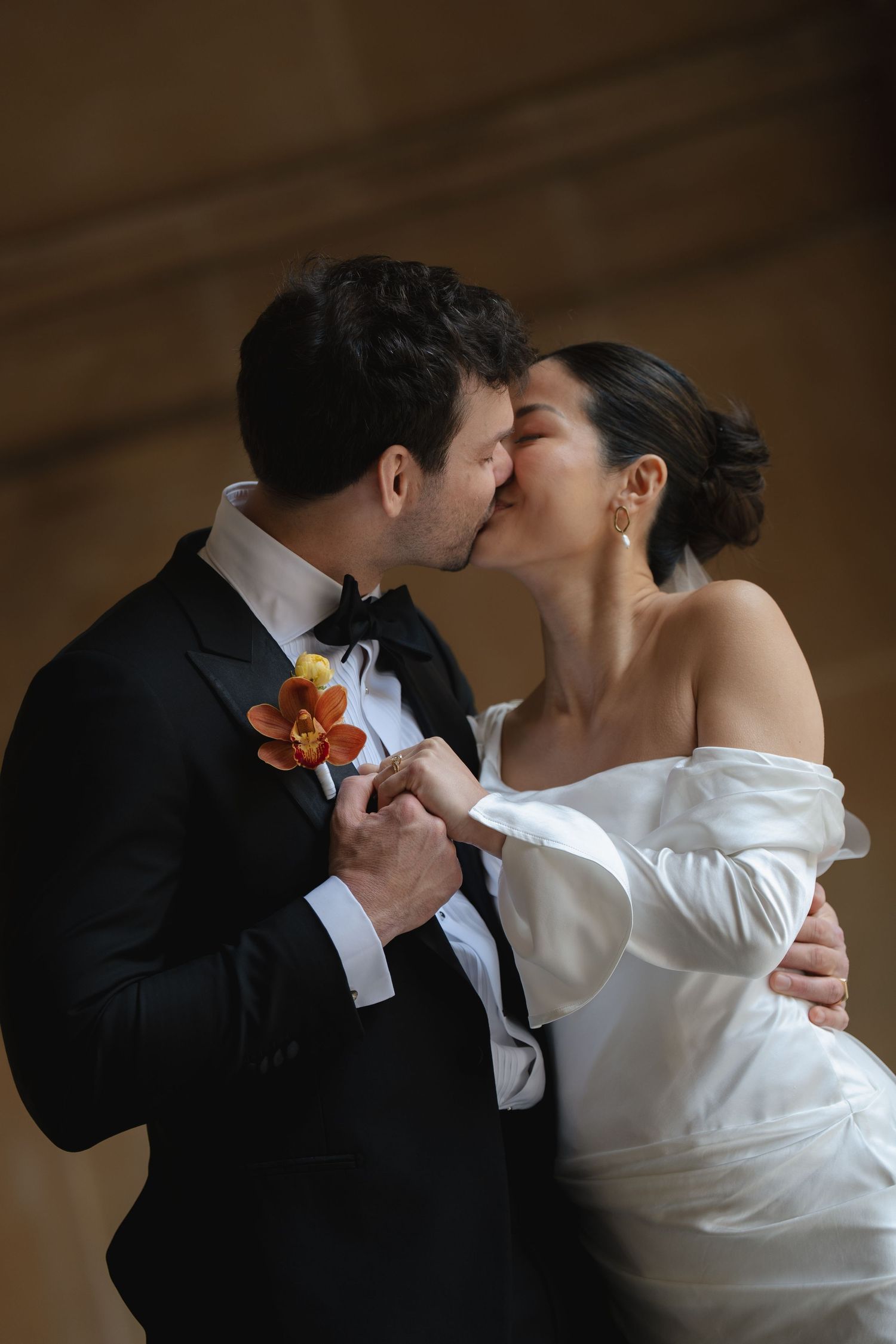 Bride and groom sharing a romantic kiss, she in an off-shoulder white gown, he in a black tuxedo with floral boutonniere.