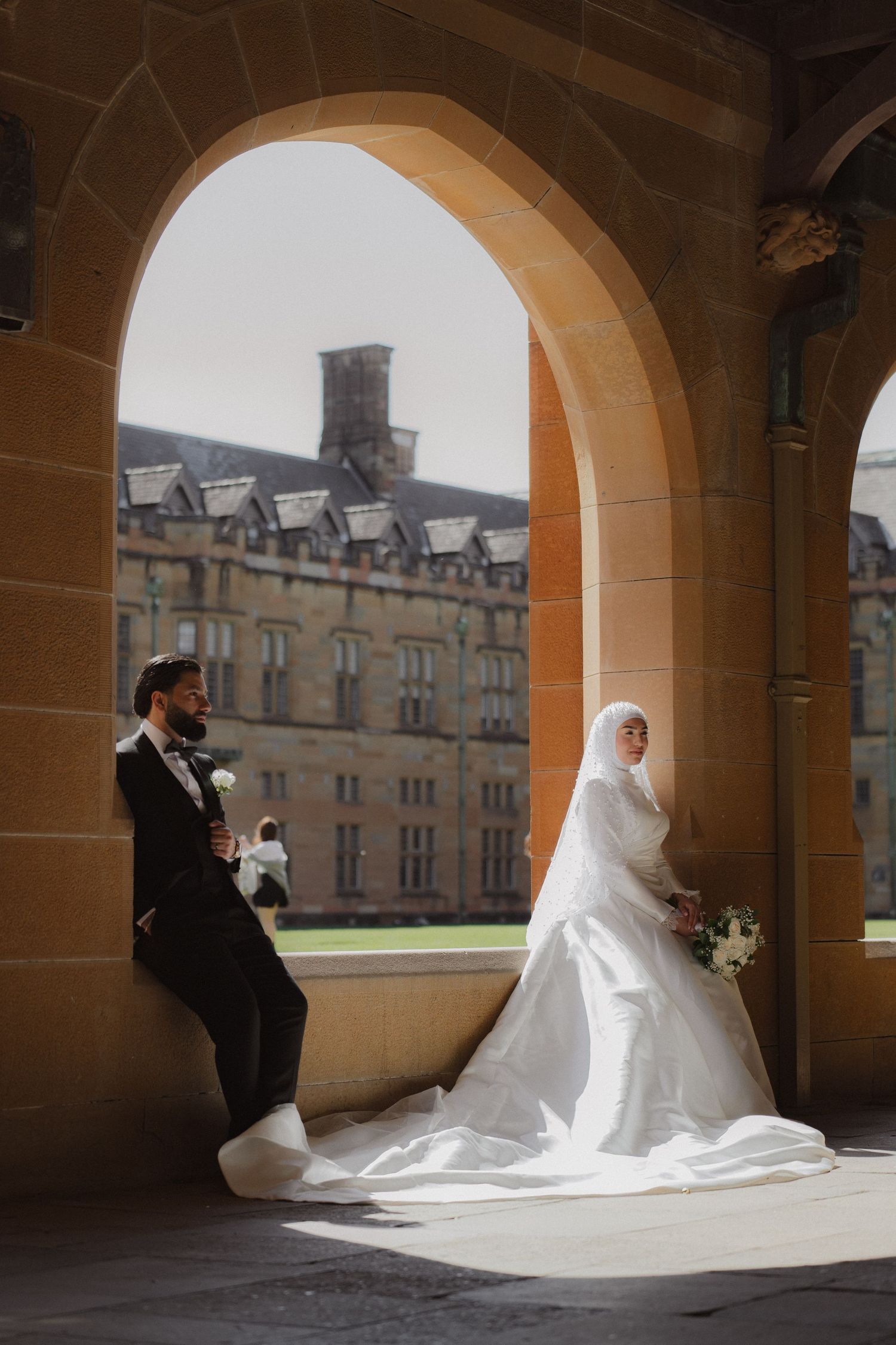 Muslim bride in white hijab and groom in black suit pose under stone arch at historic building wedding photo.