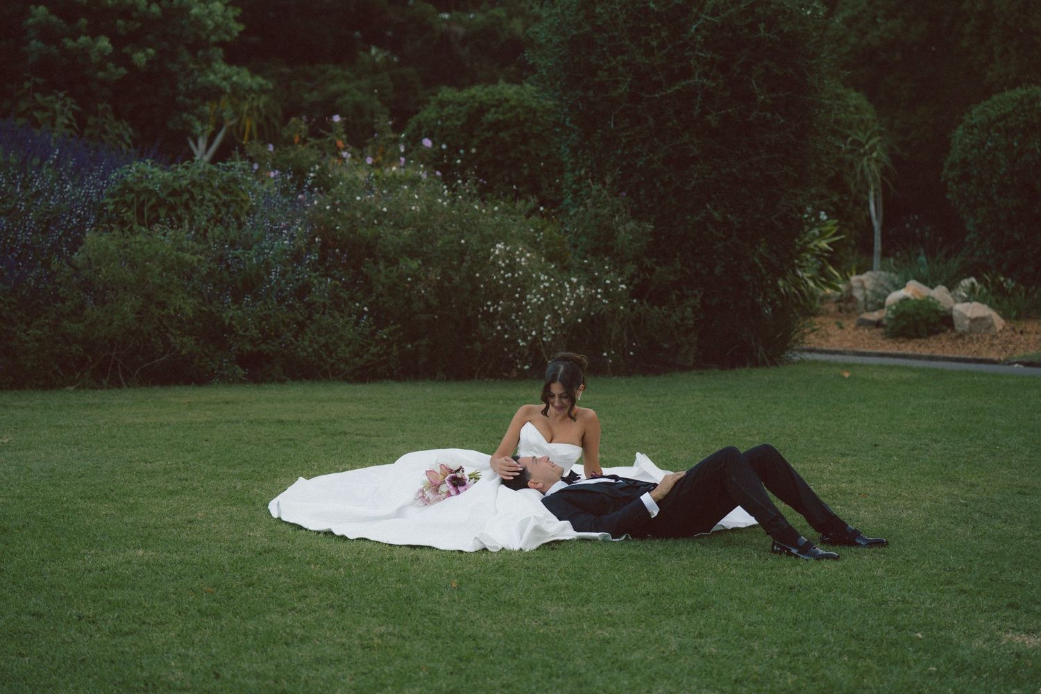 Bride in white gown and groom in black suit relaxing on green lawn in garden wedding photo.