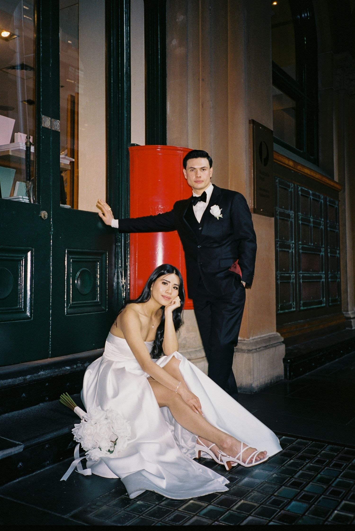 Elegant bride in white gown sits on steps while groom in black tuxedo stands beside red door at night.