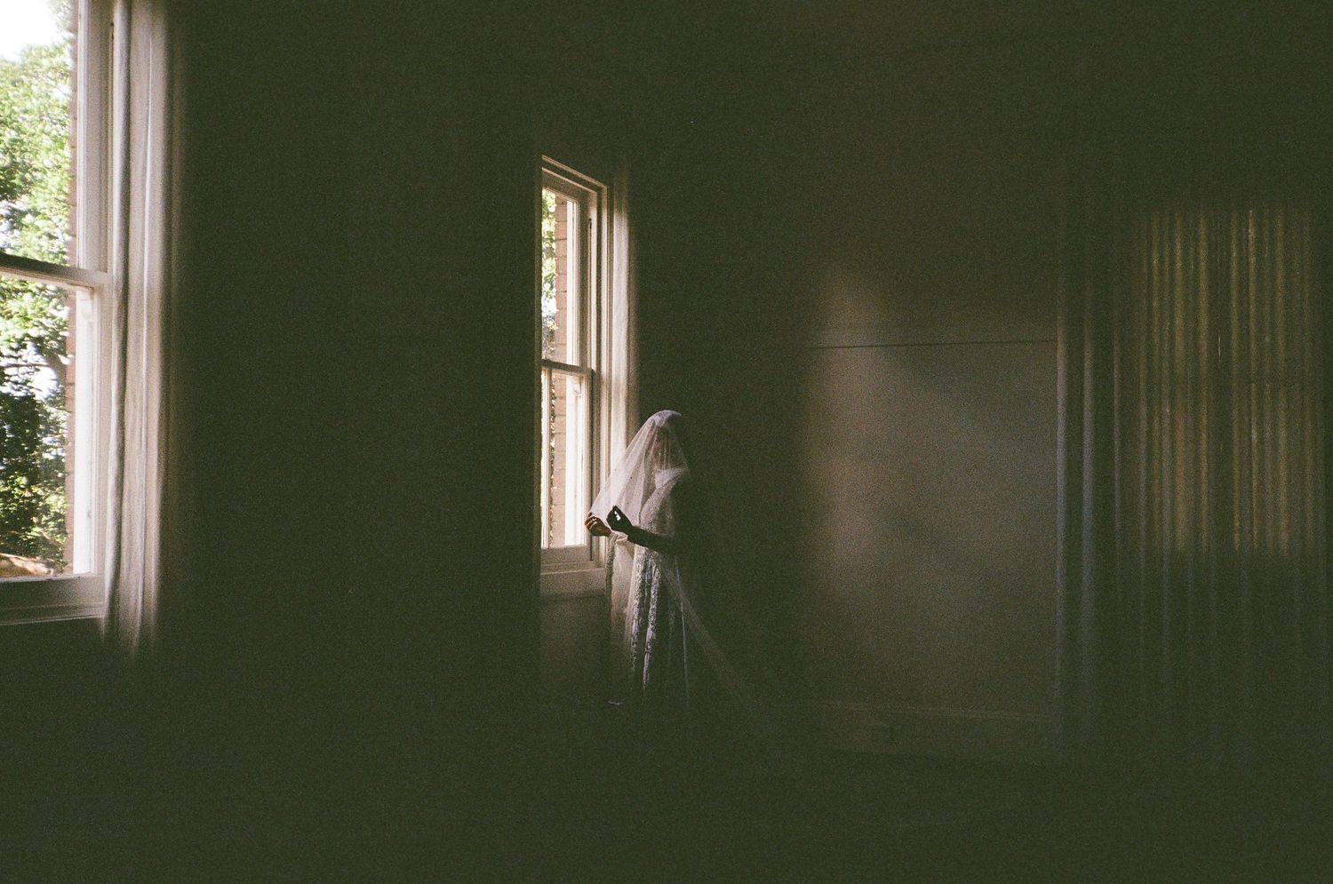 A ghostly figure in white stands by a window in a dimly lit, dark green room.