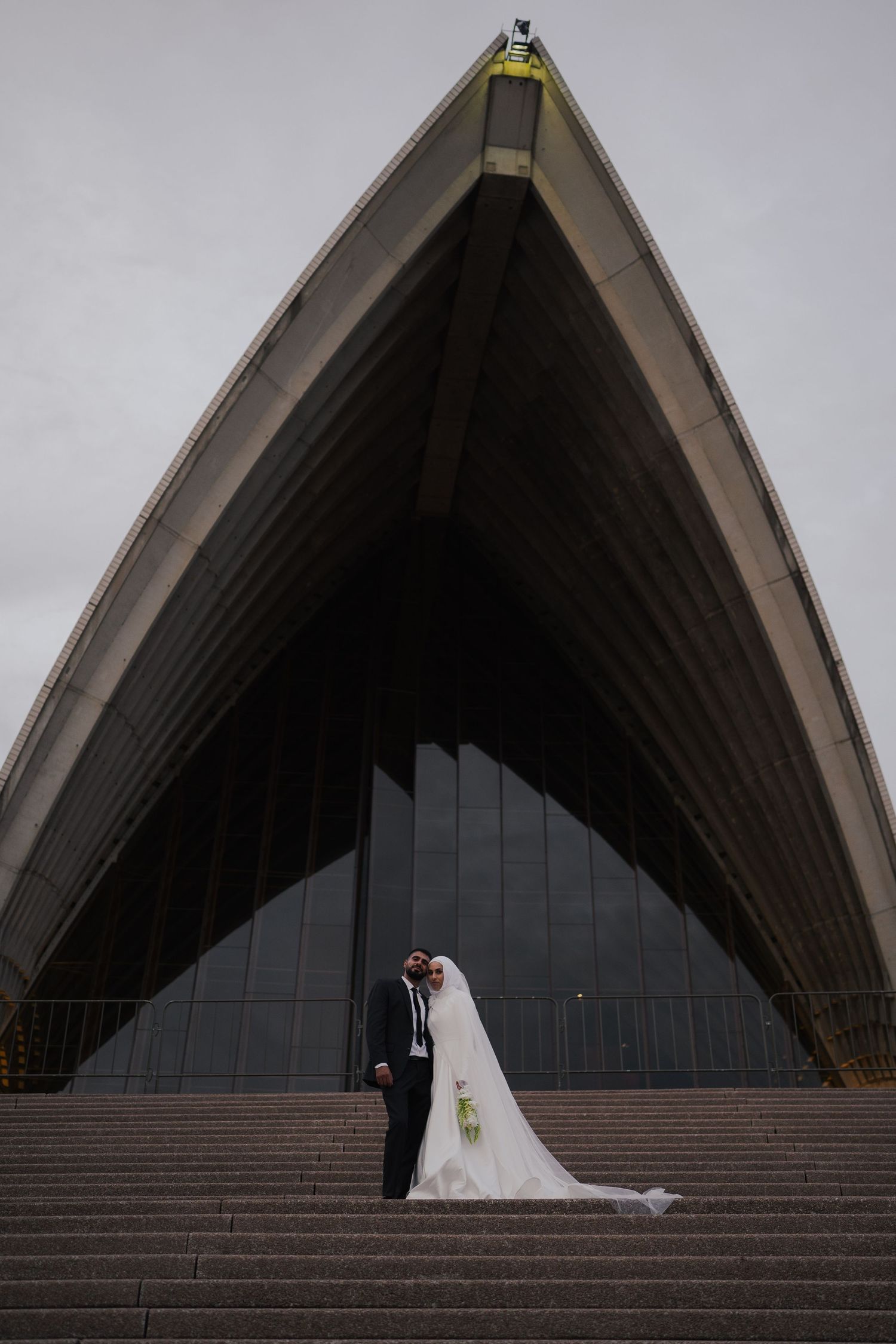 Wedding couple posing on steps of Sydney Opera House, bride in white gown with veil, groom in dark suit.