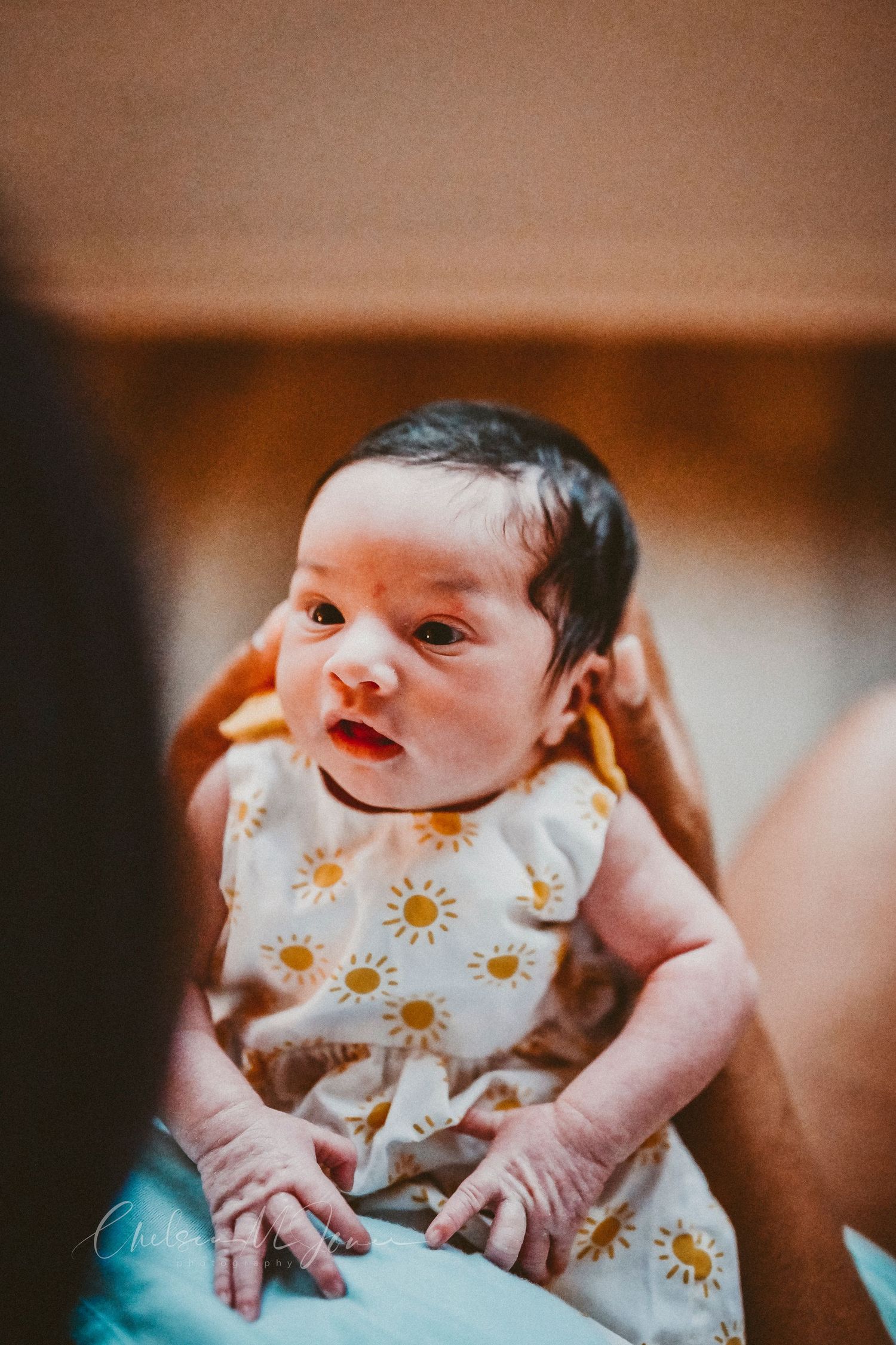 Newborn baby in a white sunflower romper being held up, looking alert with dark hair and tiny hands.