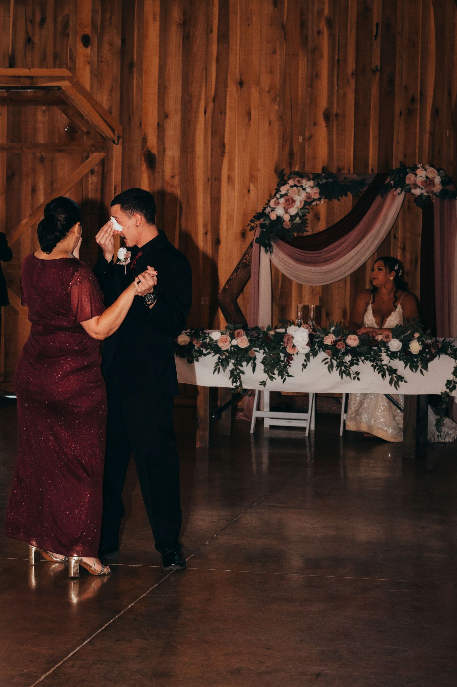 Groom shares emotional mother-son dance at rustic barn wedding reception with floral arch backdrop.
