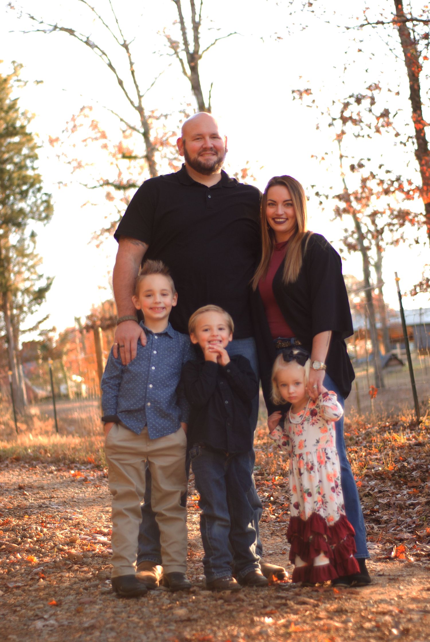 Family of five posing outdoors in autumn, parents with three young children on a leaf-covered path surrounded by fall foliage.