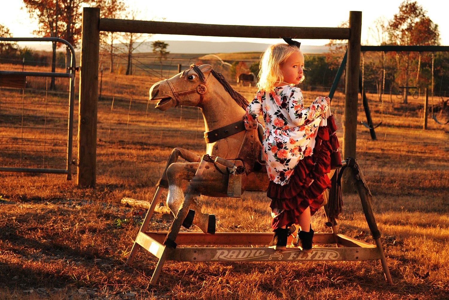 Young girl in floral dress riding a wooden rocking horse outdoors at golden hour on a rural farm.