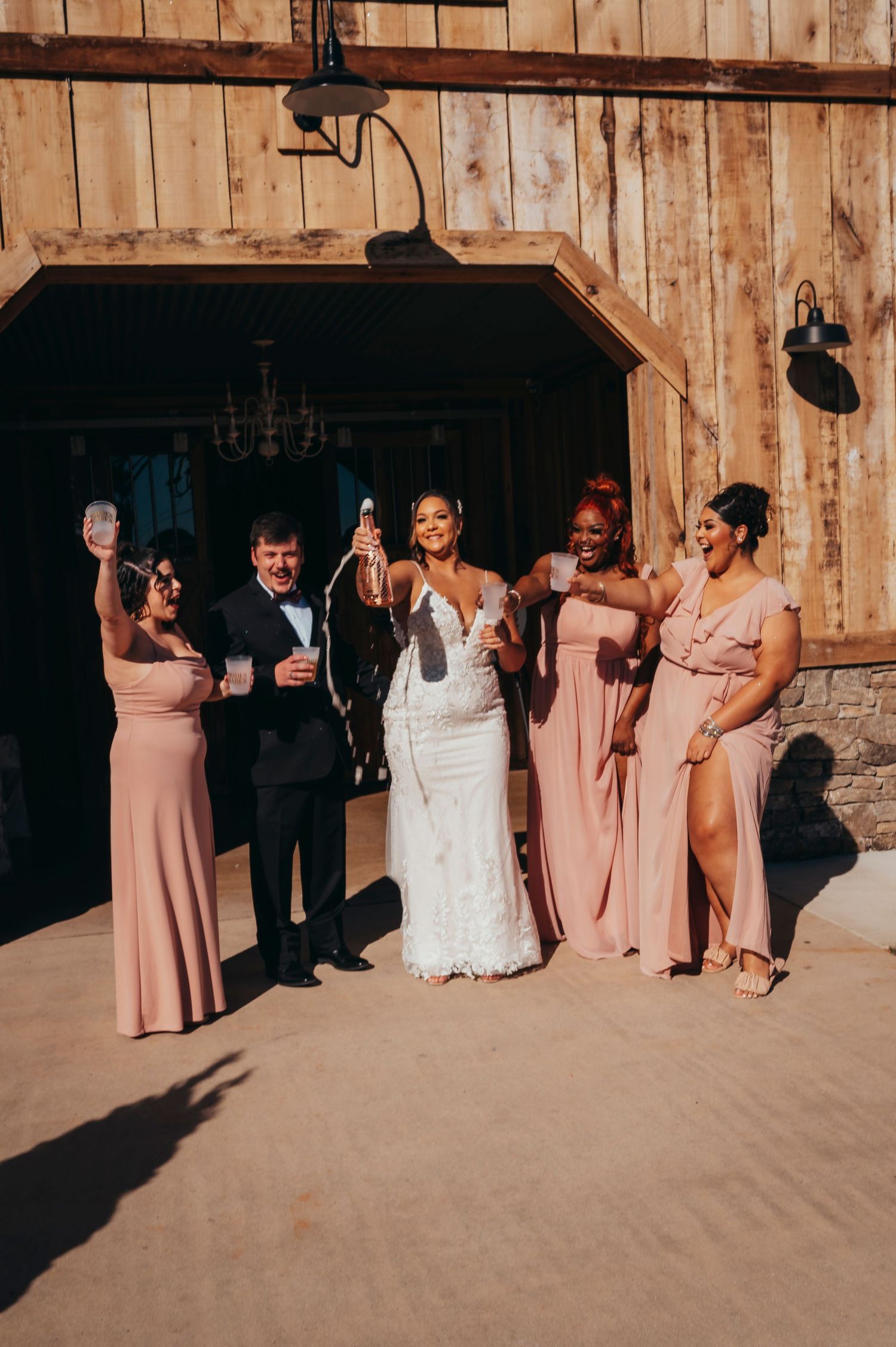 Bride in lace gown pops champagne with bridesmaids in blush pink dresses outside rustic barn wedding venue.
