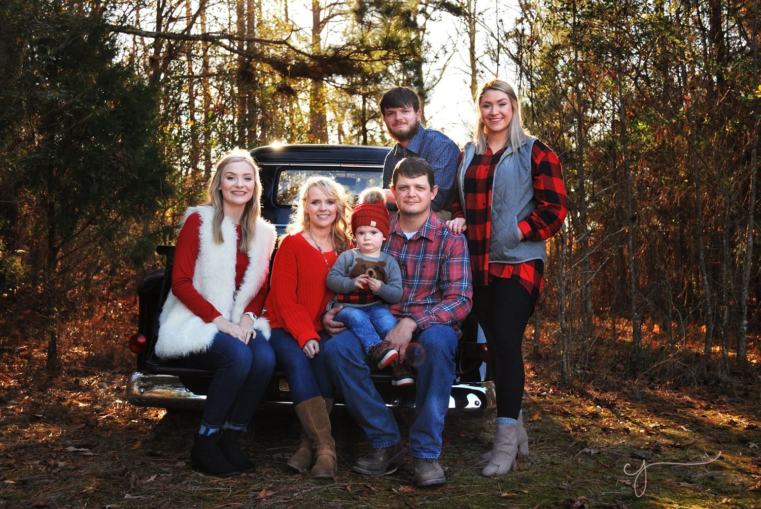 Family of six posing on vintage truck tailgate in wooded setting, wearing festive red and plaid outfits for holiday portrait.