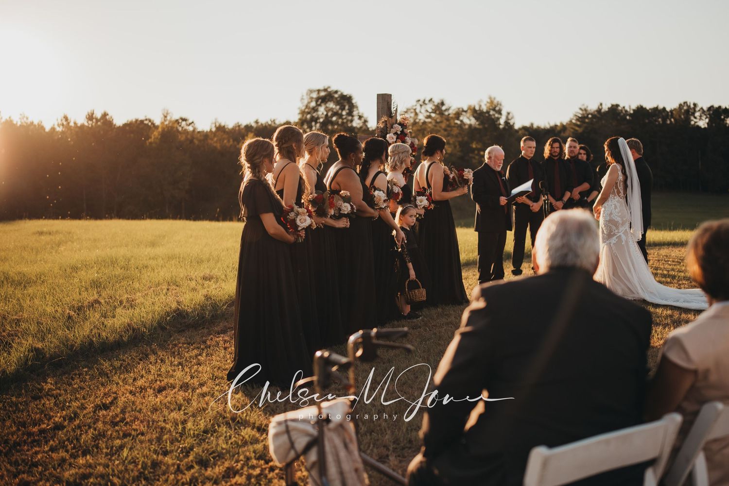 Outdoor wedding ceremony at golden hour with bridesmaids in black dresses standing beside bride in white gown on green field.