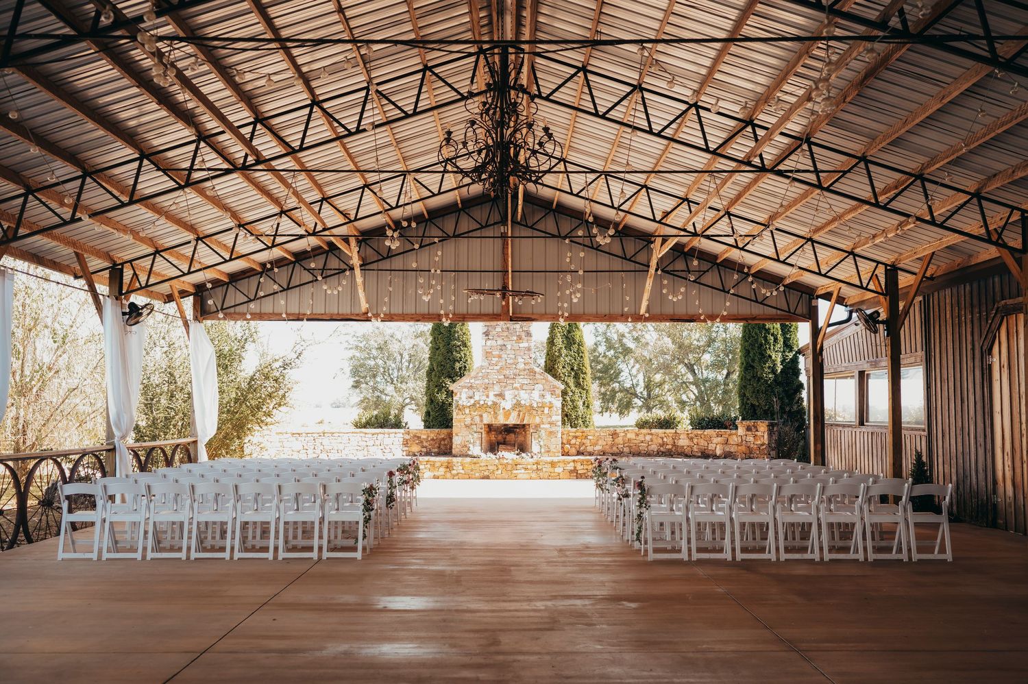 Elegant open-air barn wedding venue with white chairs arranged in rows, chandelier, and stone fireplace backdrop.