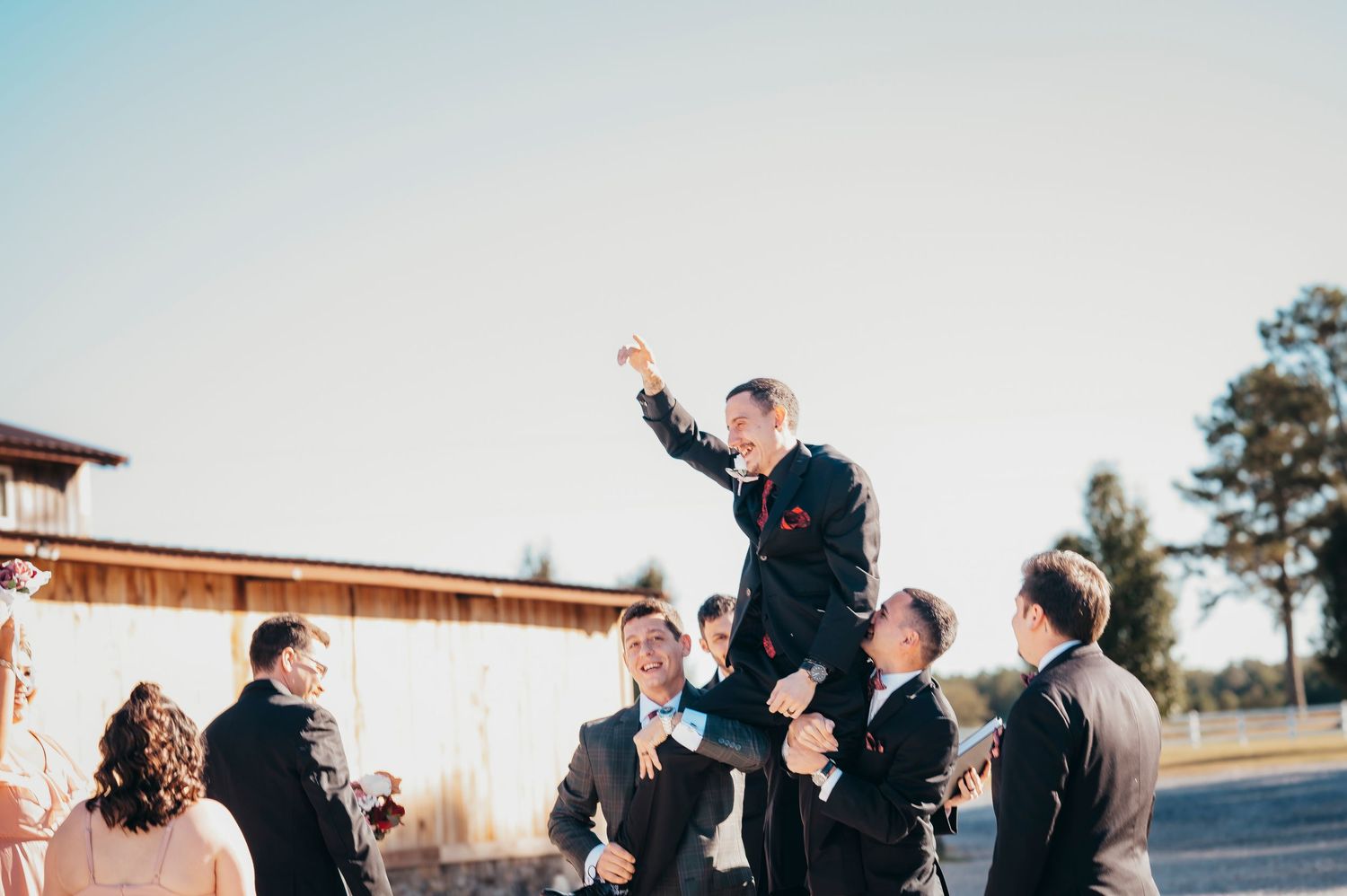 Groomsmen lifting groom in celebration outdoors at wedding venue surrounded by wedding party.