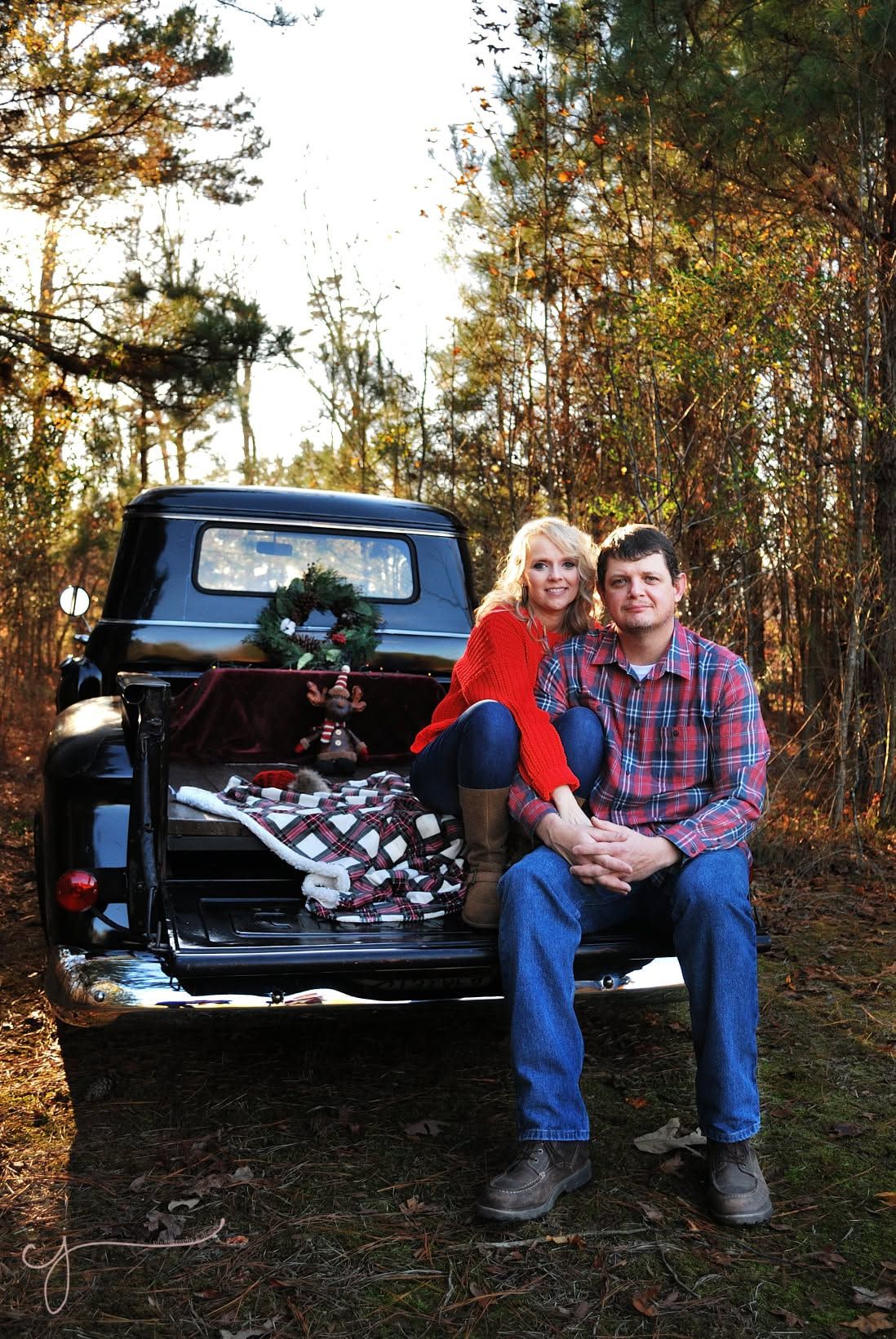 Couple sitting on vintage black truck tailgate with Christmas wreath and gifts in wooded setting.