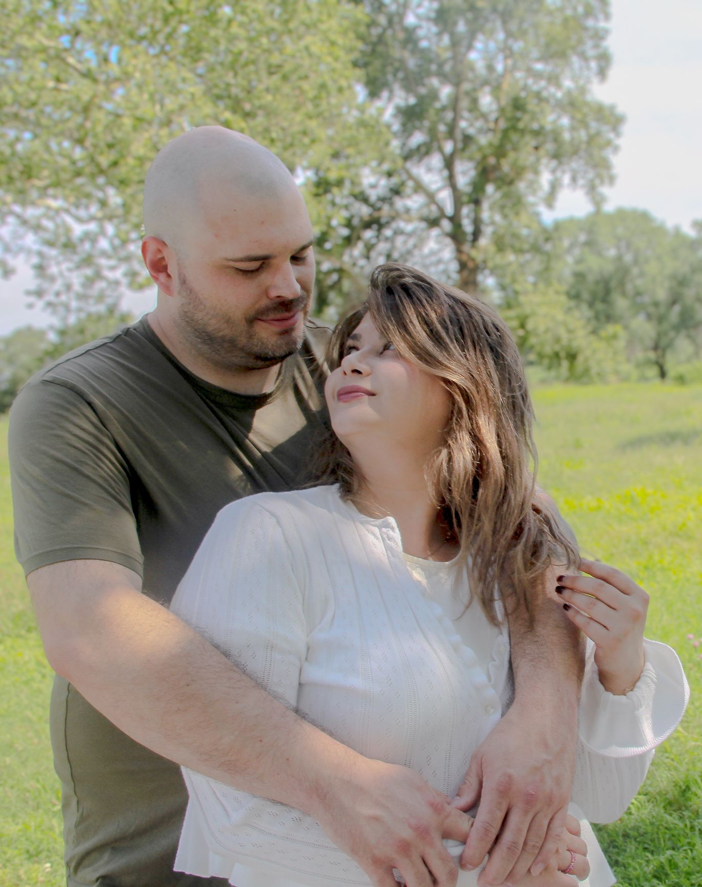 A couple embraces outdoors in a sunny green park, the man in olive shirt holding a woman in white top.