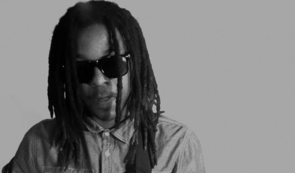 A man with dreadlocks wearing dark sunglasses poses in a moody black and white portrait photo.