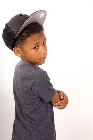 Young Black boy in gray t-shirt and black snapback cap poses with arms crossed, looking over shoulder.