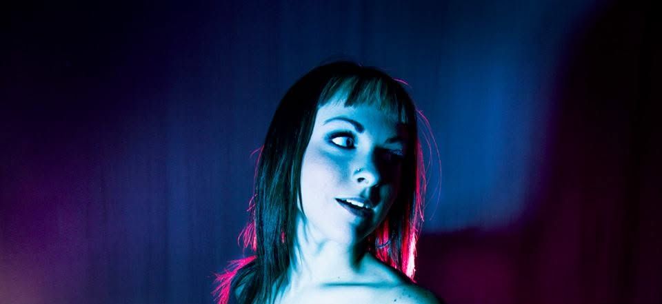 Woman with dark hair posed under dramatic blue and pink neon lighting in a moody studio portrait.
