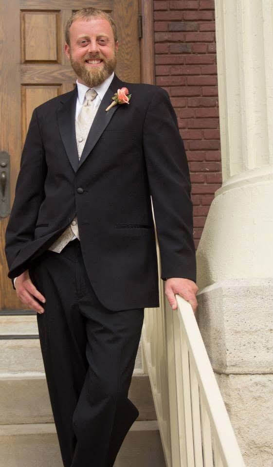 Smiling bearded man in black tuxedo with boutonniere posing on steps outside brick building.