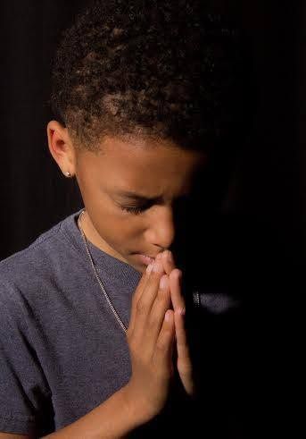 Young boy with eyes closed holds hands together in prayer pose against dark background.