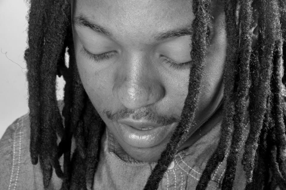 Black and white close-up of a man with long dreadlocks and a mustache, eyes closed, looking down.