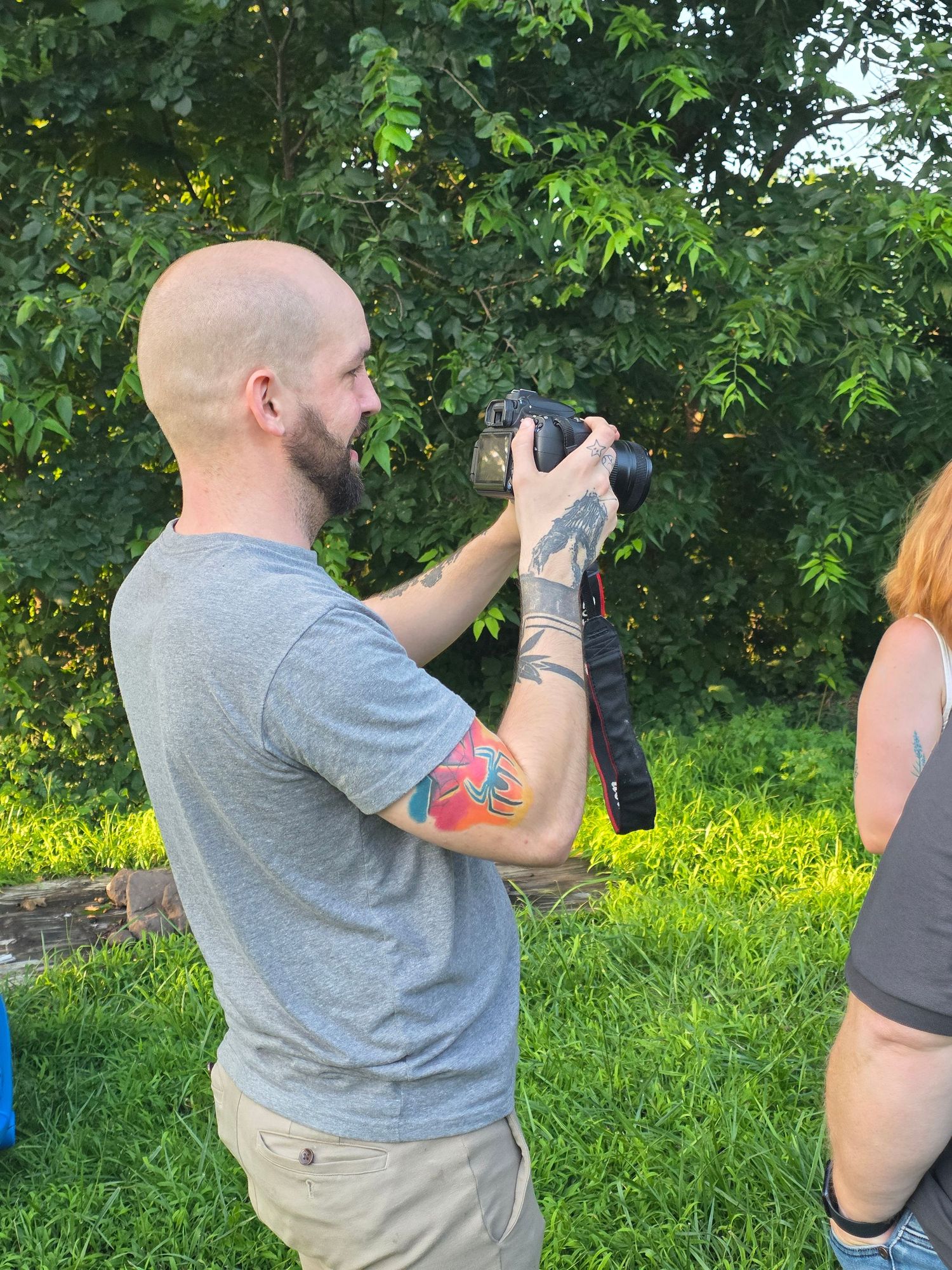 Bald man with colorful arm tattoos photographing outdoors with DSLR camera, green trees in background.