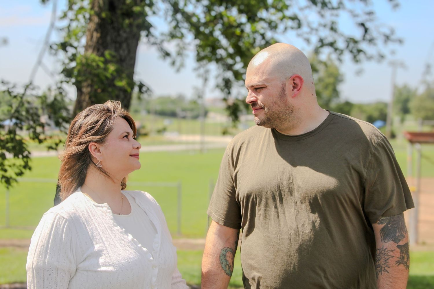 A man and woman share a warm moment outdoors, standing together near green trees on a sunny day.