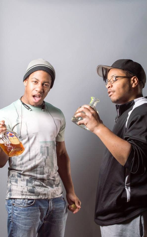 Two young men posing with liquor bottles in a studio photo shoot against a gray background.