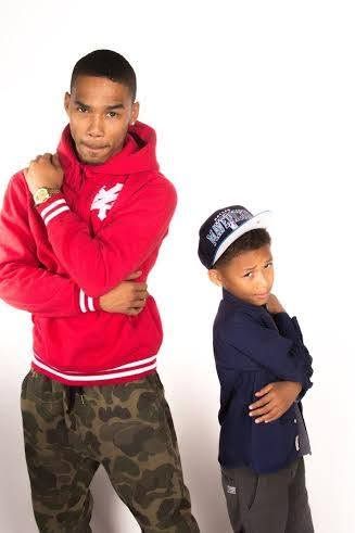 Two young males in streetwear pose with crossed arms, one in red hoodie and camo pants, other in navy shirt and snapback.