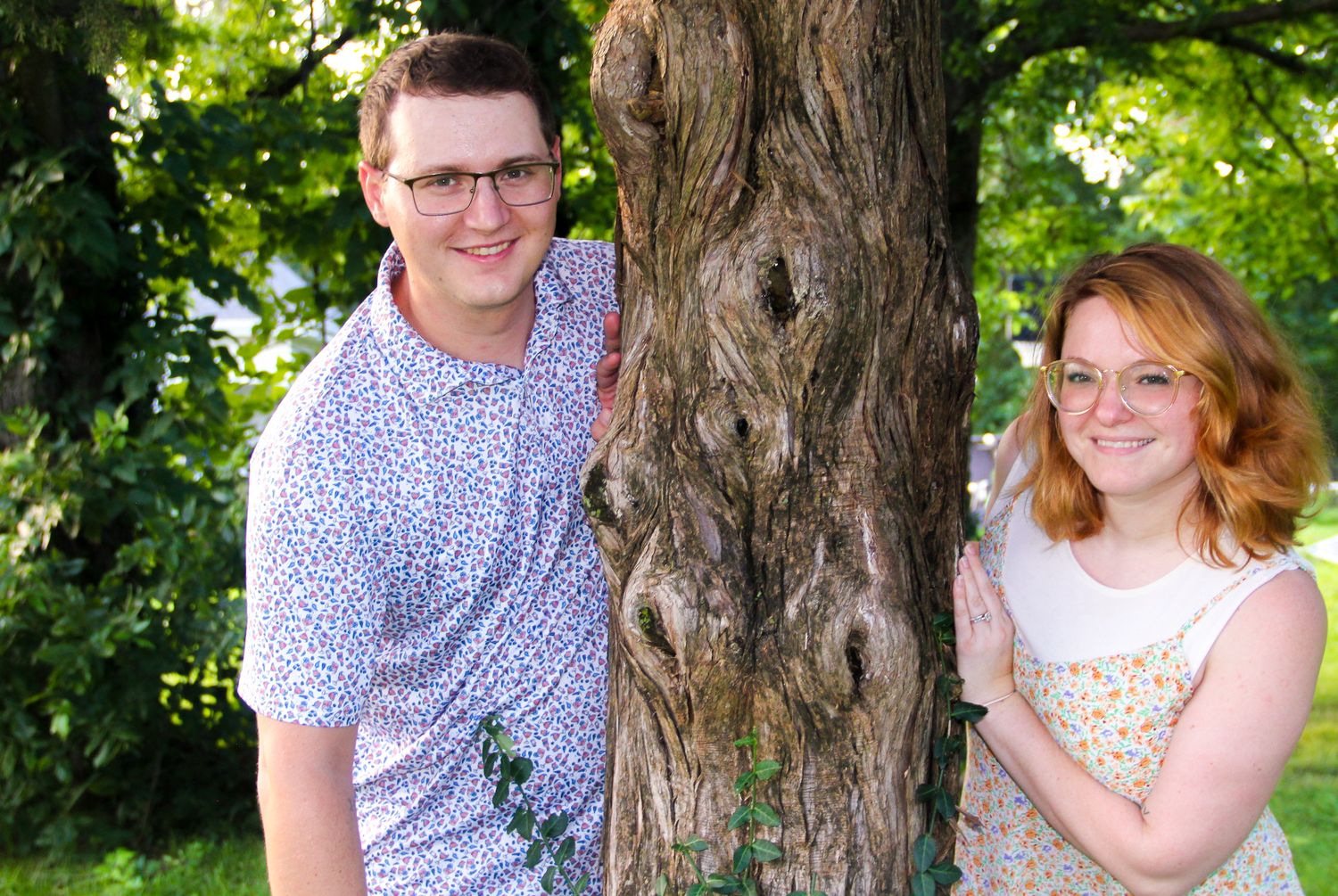A young couple smiling and posing beside a large tree trunk in a lush green park setting.