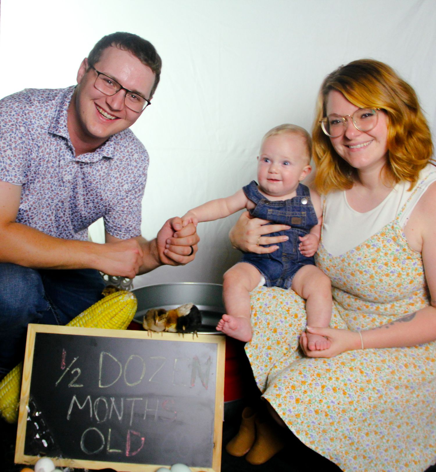 Happy family photo with parents smiling beside their 6.5 month old baby holding a chalkboard sign.