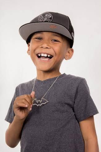 Happy young boy wearing a snapback cap and Batman necklace smiling brightly in a gray t-shirt.