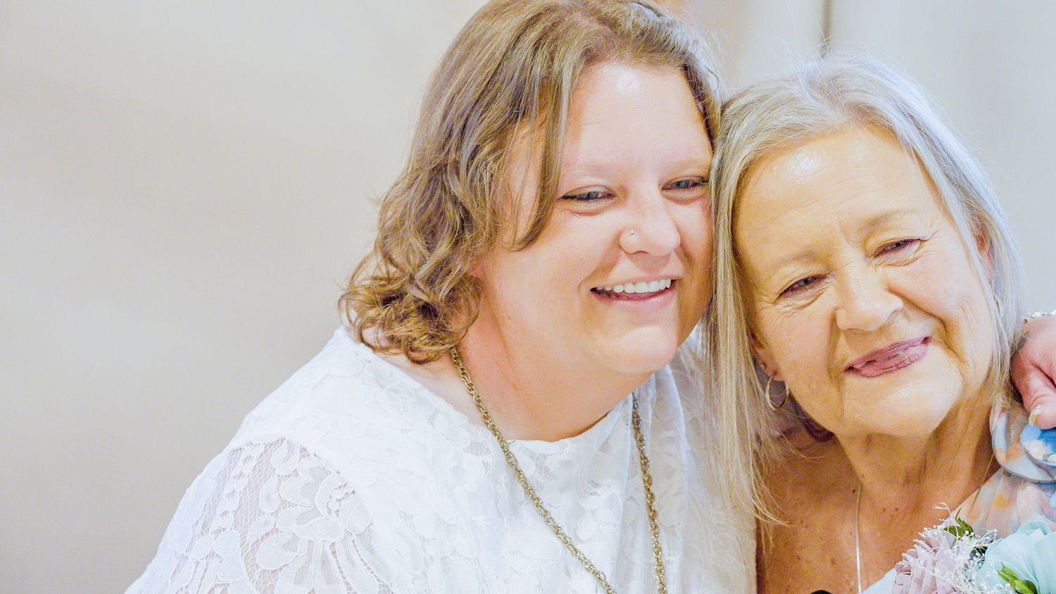 Two smiling women, one middle-aged and one elderly, pose together in white outfits for a warm portrait photo.