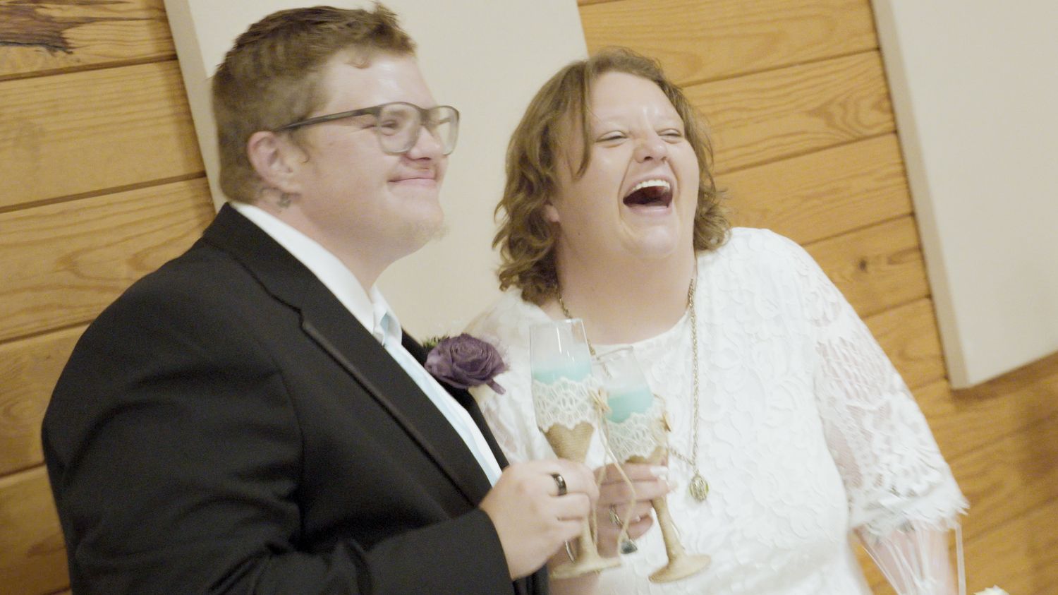 Joyful couple in wedding attire laughing together, groom in black tuxedo and bride in white lace dress.