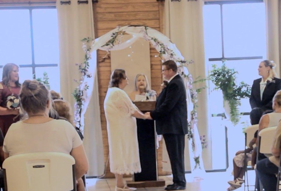 Couple exchanging vows at a rustic wedding ceremony beneath a floral arch, officiant and guests in attendance.