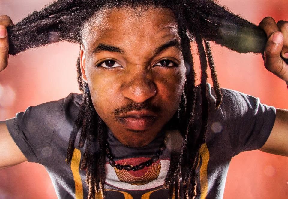 A young man with long dreadlocks pulls his hair back against a vibrant red bokeh background.