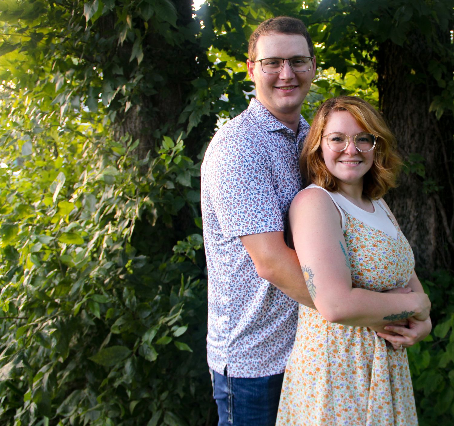 Smiling couple posing outdoors in front of lush green foliage, wearing casual summer attire.