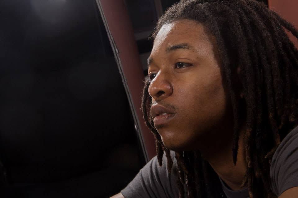Young man with dreadlocks looking intently to the side in a dimly lit setting.