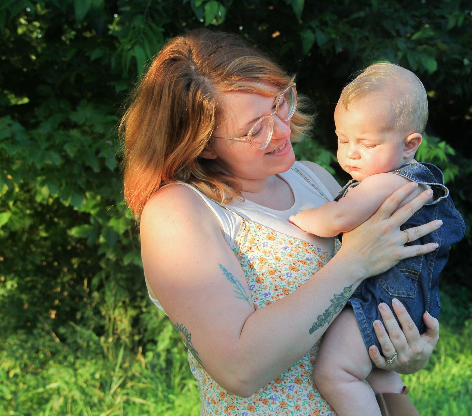 A red-haired woman in a floral dress holds a baby outdoors near green trees in warm golden sunlight.