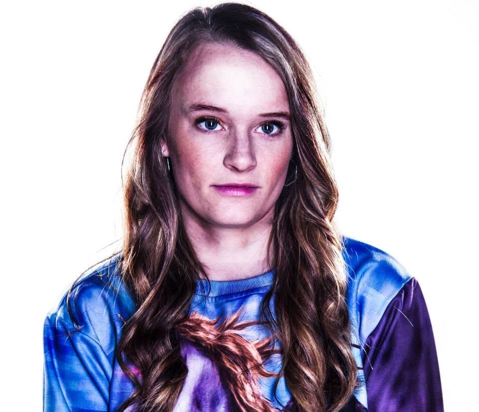 Young woman with long brown hair wearing a colorful blue graphic sweatshirt against a white background.
