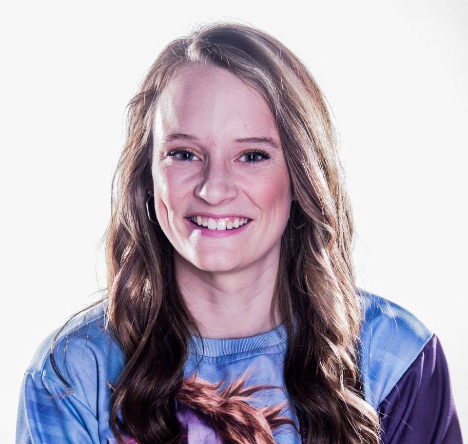 Cheerful young woman with long wavy brown hair smiling warmly wearing a blue graphic t-shirt.