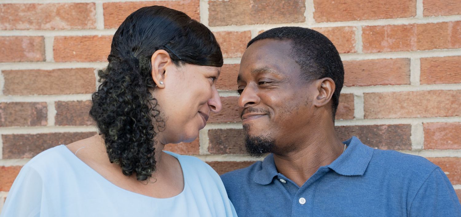 A couple smiling and gazing at each other affectionately in front of a brick wall.