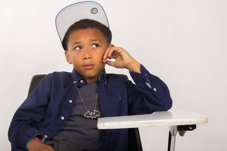Young boy in blue shirt and backwards cap sits thoughtfully in a chair, resting hand on cheek.
